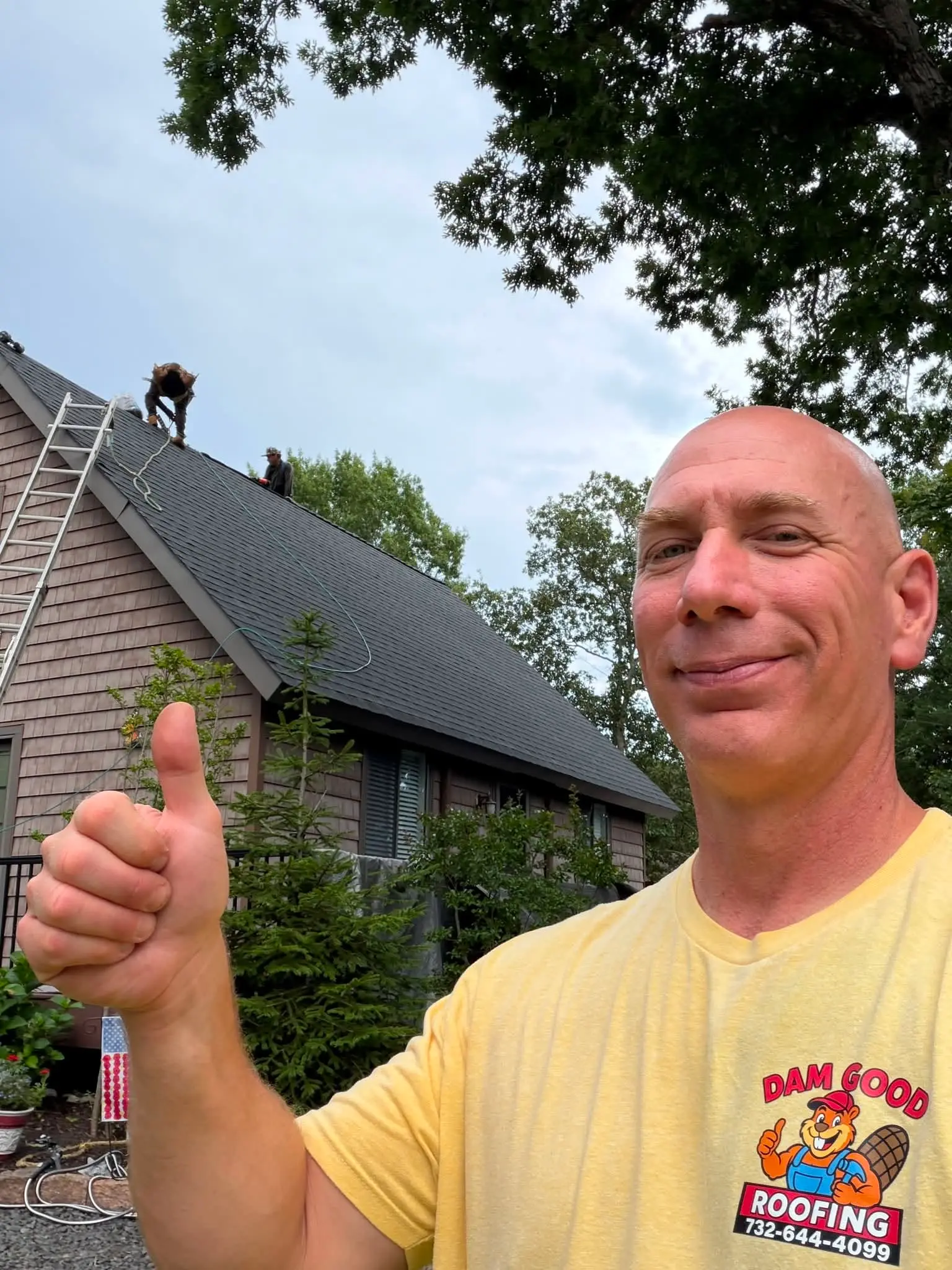 Dam Good Roofing owner in a company t-shirt giving a thumbs up while the crew replaces a roof behind him